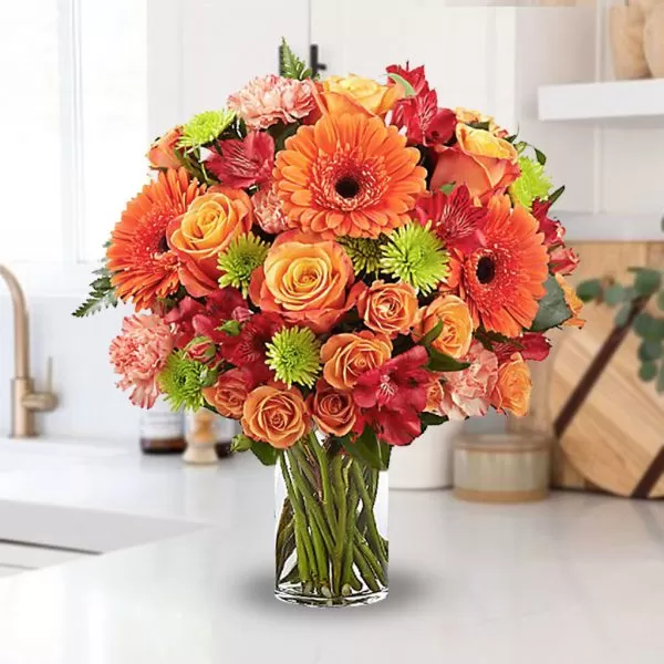 A clear glass vase filled with water and a vibrant bouquet of orange gerbera daisies, orange roses, red alstroemeria, pink carnations, and green button chrysanthemums, placed on a white surface with a blurred background. The bouquet is arranged in a dome shape, and the flowers are in full bloom.