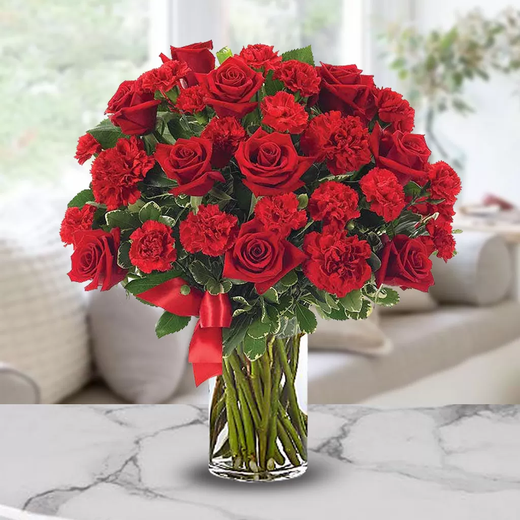 A clear glass vase filled with red roses and red carnations, tied with a red ribbon, placed on a light-colored surface with a blurred background.