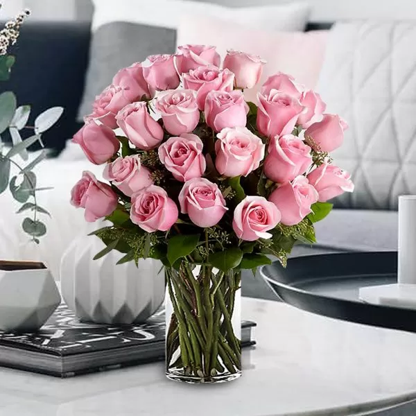 A close-up shot of a clear glass vase filled with pink roses and green stems, arranged in a tight bouquet, placed on a light-colored table with decorative items and a blurred background.