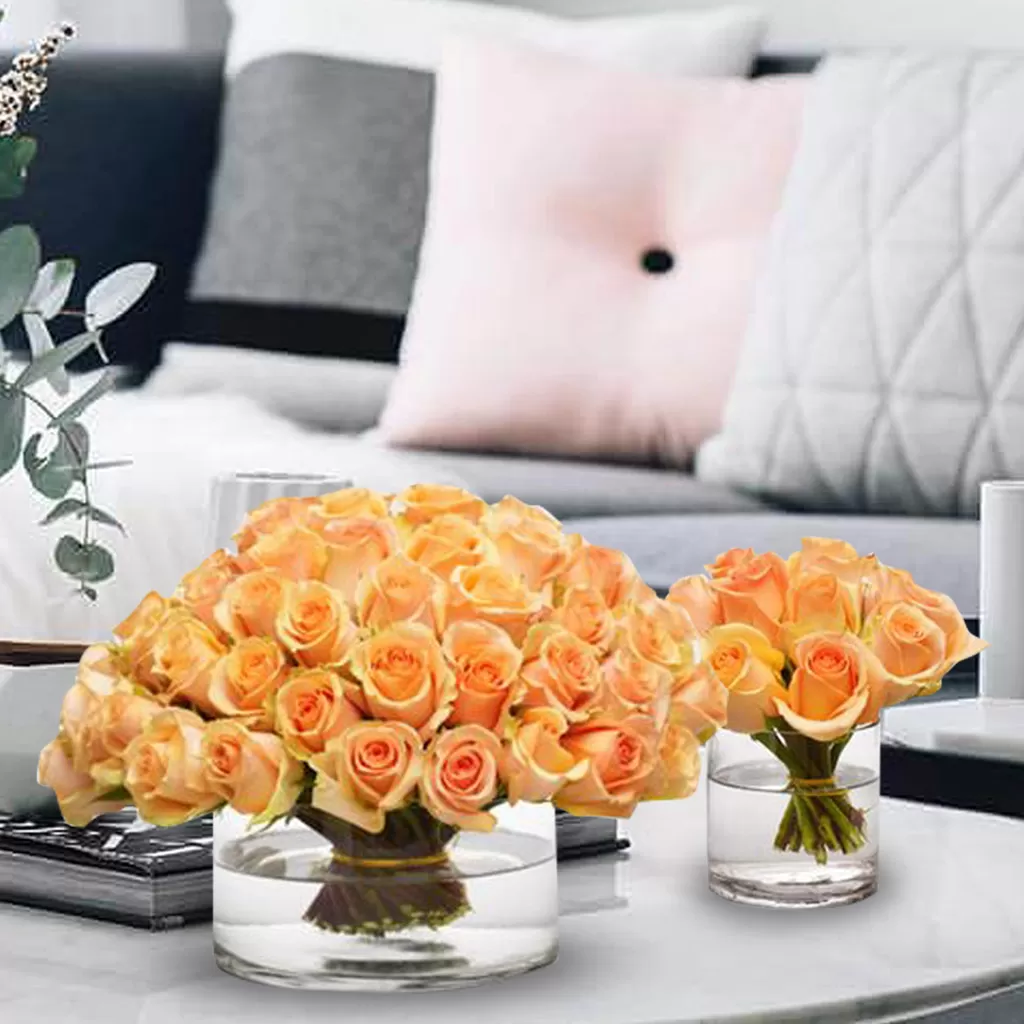 Two clear glass vases filled with water and peach-colored roses are placed on a white surface. The larger vase on the left holds a dome-shaped arrangement of numerous roses, while the smaller vase on the right contains a smaller bouquet of the same flowers. The background is a soft, blurred light with a round, bright spot.