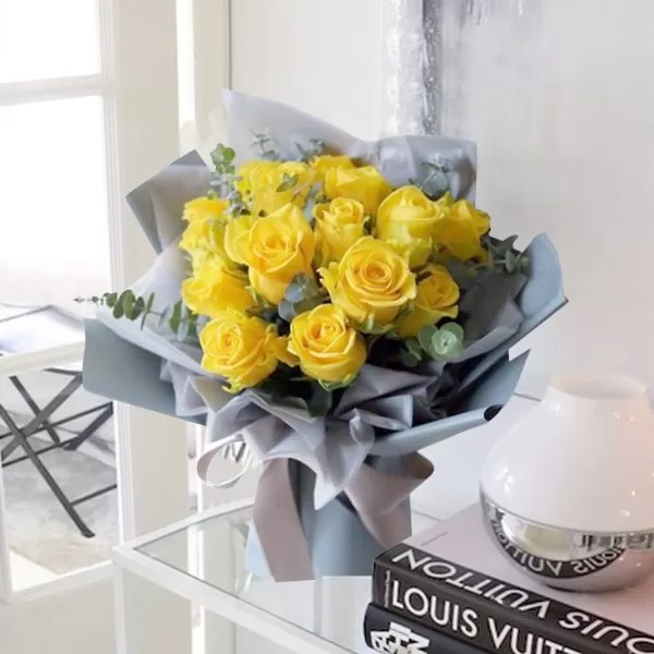 A bouquet of yellow roses with green eucalyptus leaves, wrapped in light gray paper and tied with a light gray ribbon, placed on a light wood table with a blurred background.