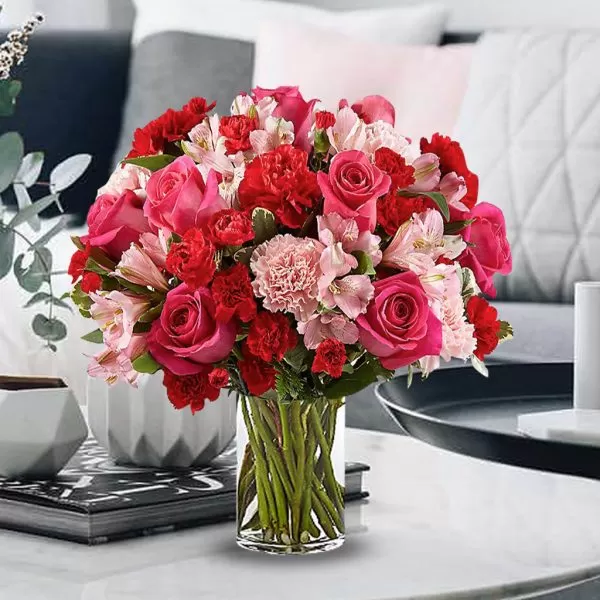 A close-up shot of a clear glass vase filled with red roses, pink roses, red carnations, pink carnations, and pink alstroemeria flowers, with visible green stems in the water, placed on a light-colored surface with a blurred background.