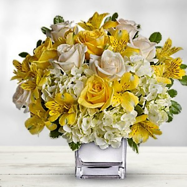 A square, clear glass vase filled with yellow alstroemeria, yellow roses, white roses, and white hydrangeas, placed on a white surface.