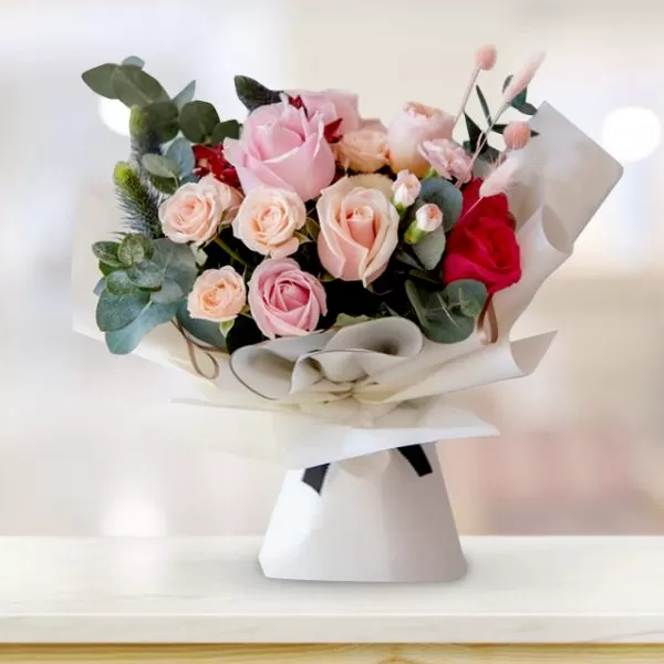 A close-up shot of a bouquet featuring pink roses, red roses, and greenery (possibly eucalyptus), wrapped in white paper with a black tie, placed on a light-colored surface with a blurred background.