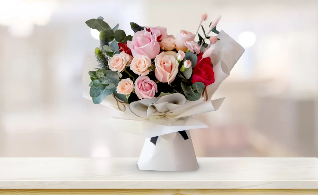 A close-up shot of a bouquet featuring pink roses, red roses, and greenery (possibly eucalyptus), wrapped in white paper with a black tie, placed on a light-colored surface with a blurred background.