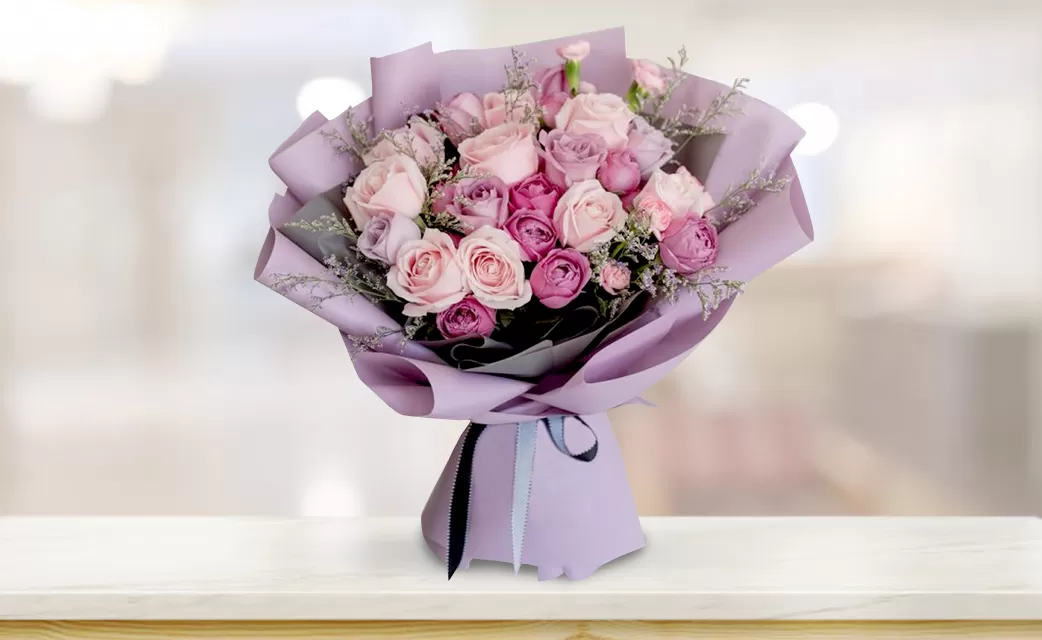 A bouquet featuring pink and deep purple roses, accented with small sprigs of greenery, wrapped in light purple paper and tied with a black ribbon, displayed against a blurred background.