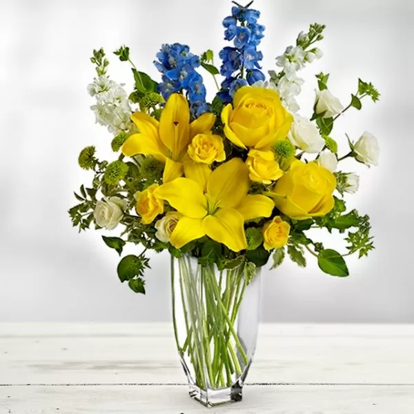 A clear glass vase filled with water and a bouquet of yellow lilies, yellow roses, blue delphiniums, and white snapdragons, placed on a white wooden surface. The flowers are arranged in a classic style, with the lilies as the focal point.