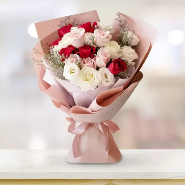 A bouquet of red, pink, and white roses wrapped in light pink paper and tied with a light pink ribbon, placed on a light-colored surface with a blurred background.