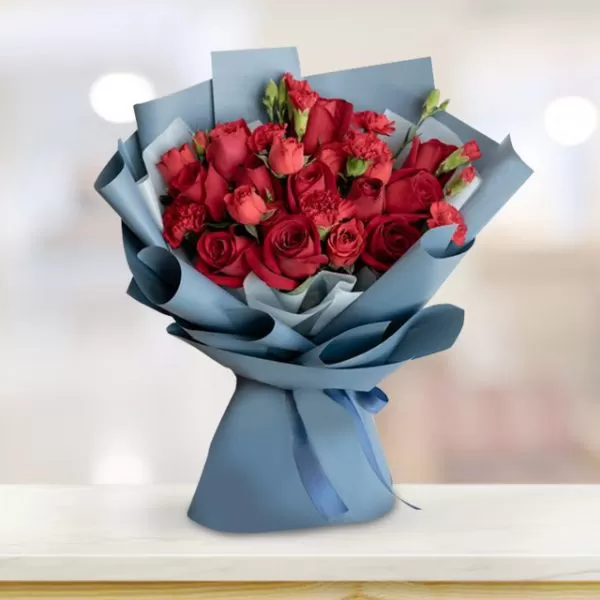 A bouquet of red roses and red carnations wrapped in light blue paper and tied with a light blue ribbon, placed on a light-colored surface with a blurred background.