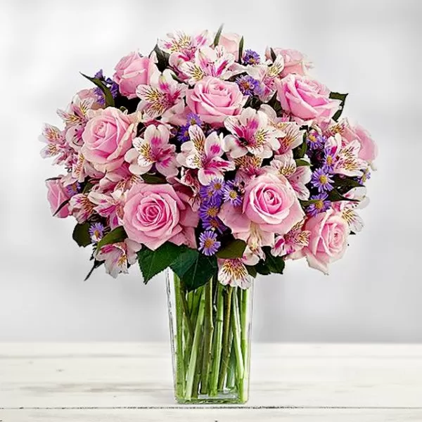 A clear glass vase filled with water and a bouquet of pink roses and pink alstroemeria, placed on a white wooden surface. The flowers are arranged in a dome shape, with the roses as the focal point. The background is a soft, blurred light.