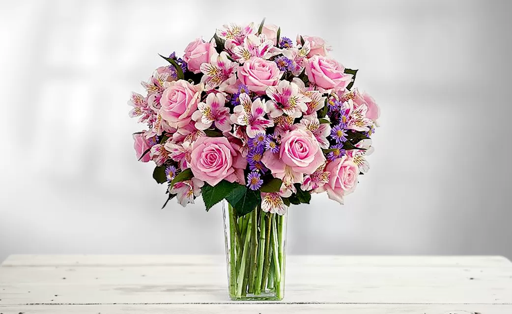 A clear glass vase filled with water and a bouquet of pink roses and pink alstroemeria, placed on a white wooden surface. The flowers are arranged in a dome shape, with the roses as the focal point. The background is a soft, blurred light.