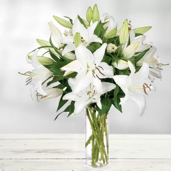 The image shows a clear glass vase filled with water and a bouquet of white lilies, placed on a white wooden surface. The lilies are in full bloom and have a fresh, vibrant appearance. The background is a soft, blurred light.