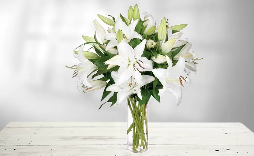 The image shows a clear glass vase filled with water and a bouquet of white lilies, placed on a white wooden surface. The lilies are in full bloom and have a fresh, vibrant appearance. The background is a soft, blurred light.