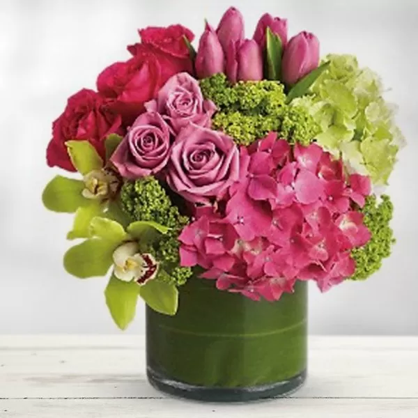 A green glass vase filled with water and a colorful bouquet of pink roses, pink tulips, pink hydrangeas, green cymbidium orchids, and green button chrysanthemums, placed on a white wooden surface. The flowers are arranged in a compact, dome-shaped design, with the hydrangeas as the focal point.