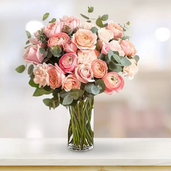 A close-up shot of a clear glass vase filled with pink and peach roses and green eucalyptus leaves, arranged in a tight bouquet, placed on a light-colored surface with a blurred background.