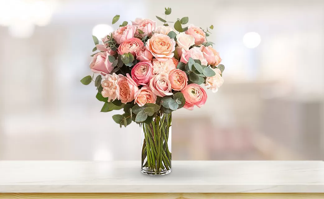 A close-up shot of a clear glass vase filled with pink and peach roses and green eucalyptus leaves, arranged in a tight bouquet, placed on a light-colored surface with a blurred background.