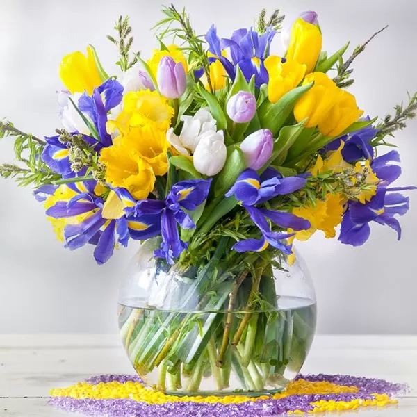 A clear, round glass vase filled with yellow daffodils, yellow and purple tulips, purple irises, and white flowers, placed on a white surface with yellow and purple decorative crystals scattered around the base.