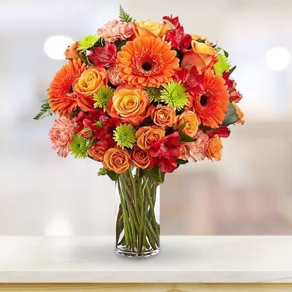 A clear glass vase filled with water and a vibrant bouquet of orange gerbera daisies, orange roses, red alstroemeria, pink carnations, and green button chrysanthemums, placed on a white surface with a blurred background. The bouquet is arranged in a dome shape, and the flowers are in full bloom.