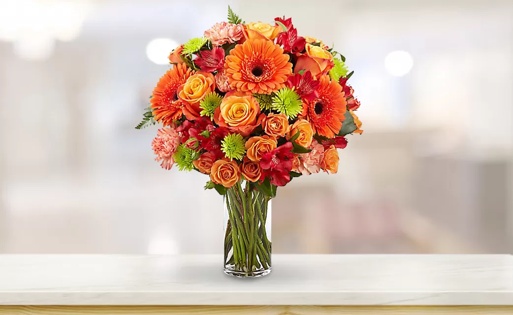 A clear glass vase filled with water and a vibrant bouquet of orange gerbera daisies, orange roses, red alstroemeria, pink carnations, and green button chrysanthemums, placed on a white surface with a blurred background. The bouquet is arranged in a dome shape, and the flowers are in full bloom.