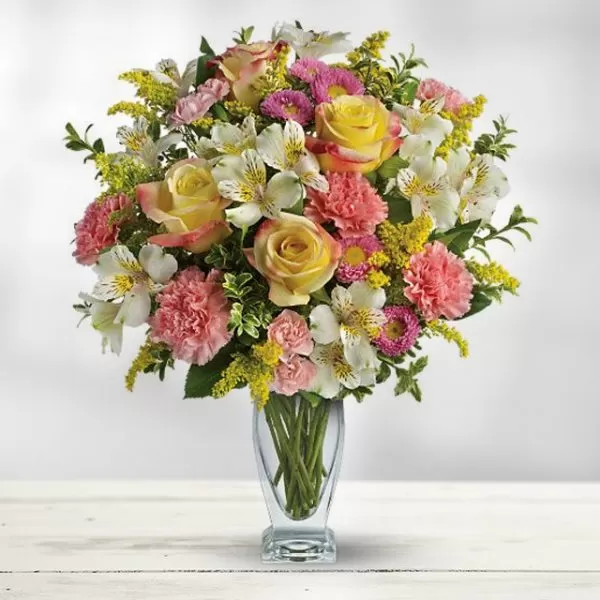 A clear glass vase filled with water and a colorful bouquet of yellow roses, pink carnations, white alstroemeria, yellow solidago, and pink asters, placed on a white wooden surface. The flowers are arranged in a dome shape, with the roses as the focal point.