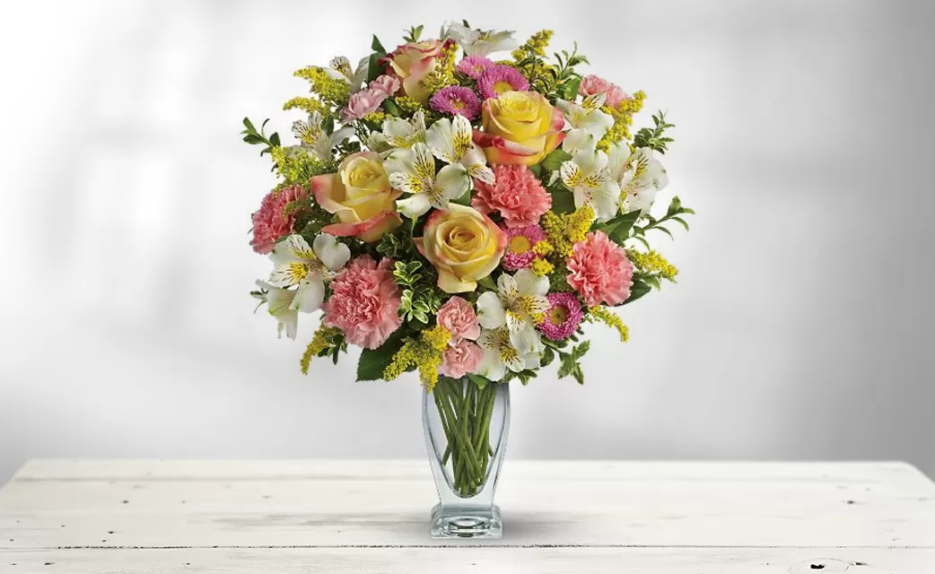 A clear glass vase filled with water and a colorful bouquet of yellow roses, pink carnations, white alstroemeria, yellow solidago, and pink asters, placed on a white wooden surface. The flowers are arranged in a dome shape, with the roses as the focal point.