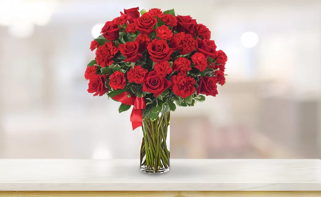 A clear glass vase filled with red roses and red carnations, tied with a red ribbon, placed on a light-colored surface with a blurred background.