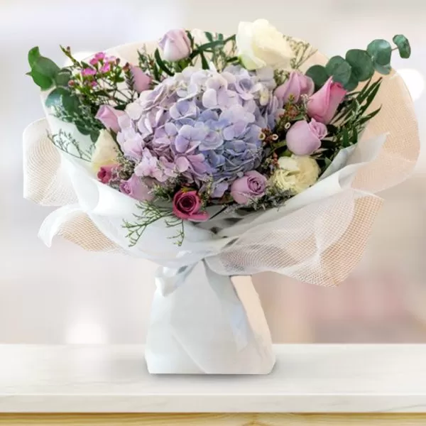 A bouquet of purple hydrangeas, pink roses, white roses, purple waxflowers, and green eucalyptus leaves, wrapped in white paper and tied with a white ribbon, placed on a light wood table with a blurred background. The bouquet is arranged in a classic style, with the hydrangeas as the focal point.