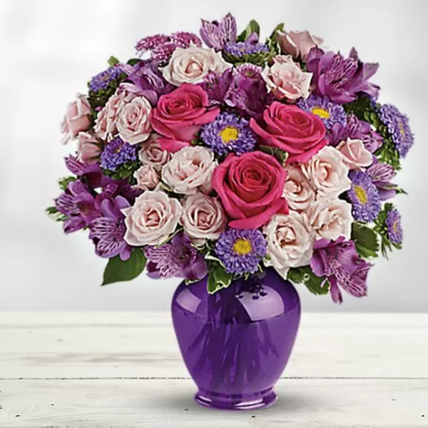 This image shows a bouquet of flowers in a purple vase. The flowers are a mix of pink and purple roses, purple alstroemeria, and purple asters. The bouquet is arranged in a dome shape and is placed on a white wooden surface. The background is a soft, blurred light.