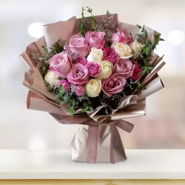 A bouquet of pink, white, and burgundy roses with green eucalyptus leaves, wrapped in light brown paper and tied with a light brown ribbon, placed on a light wood table with a blurred background. The background shows a room with a window.