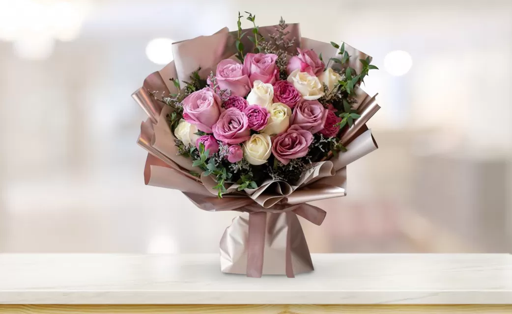 A bouquet of pink, white, and burgundy roses with green eucalyptus leaves, wrapped in light brown paper and tied with a light brown ribbon, placed on a light wood table with a blurred background. The background shows a room with a window.