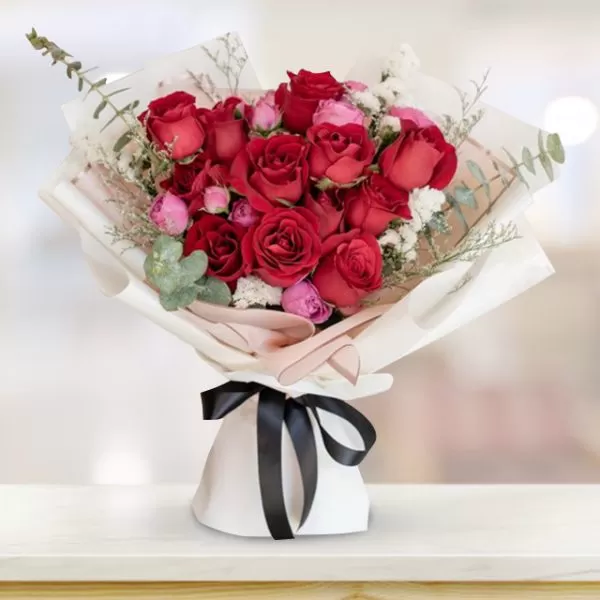 A bouquet of red roses and small pink flowers, wrapped in white paper and tied with a black ribbon, placed on a light-colored surface with a blurred background.