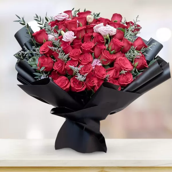 A large bouquet of red roses with a few small pink flowers, wrapped in black paper, placed on a light-colored surface with a blurred background.