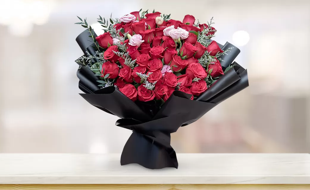 scarlet_love A large bouquet of red roses with a few small pink flowers, wrapped in black paper, placed on a light-colored surface with a blurred background.