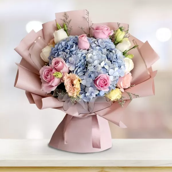 A bouquet featuring blue hydrangeas, pink and white roses, and peach eustoma flowers, wrapped in pink paper and tied with a pink ribbon, placed on a light-colored surface.