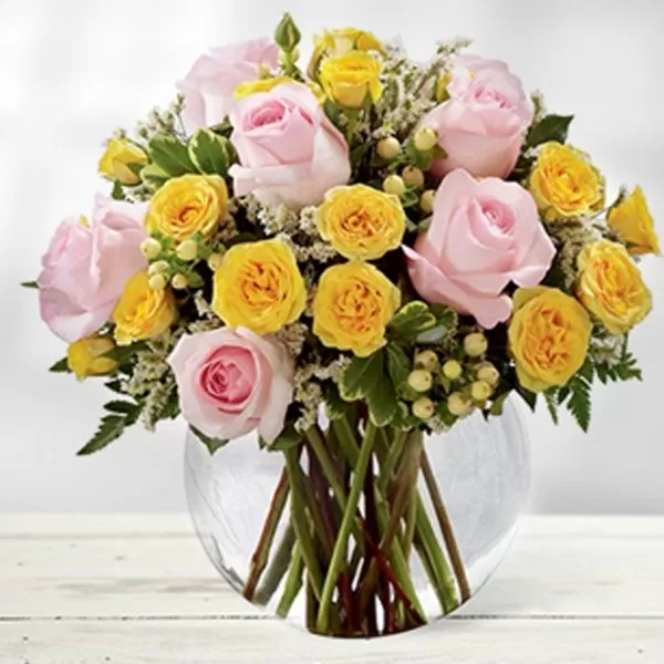 A clear glass vase filled with water and a bouquet of pink roses and yellow roses, placed on a white wooden surface. The flowers are arranged in a dome shape, with the pink roses as the focal point. The background is a soft, blurred light.