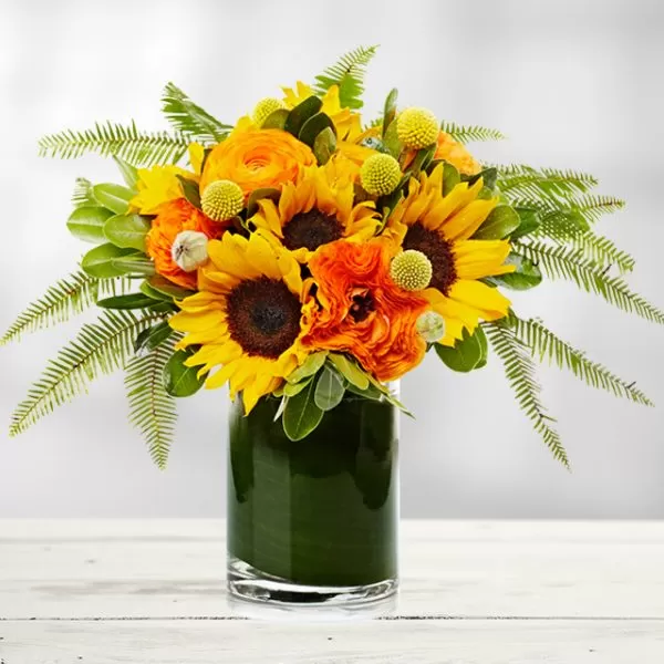 A dark green, cylindrical vase filled with yellow sunflowers, orange ranunculus flowers, yellow billy balls, and green fern leaves, placed on a white wooden table.