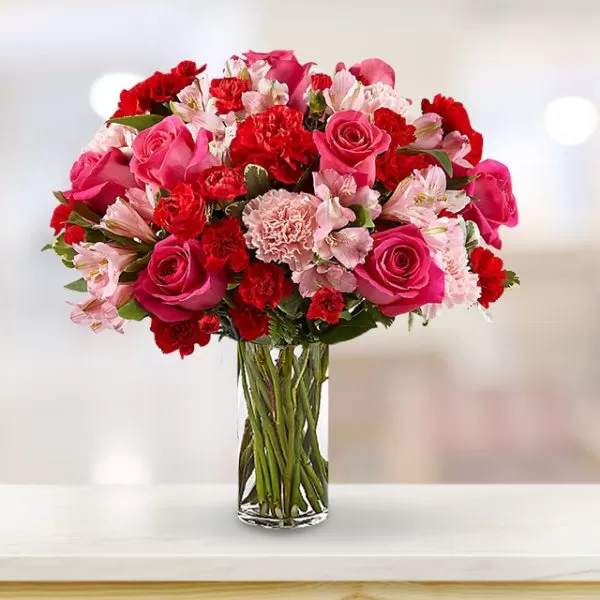 A clear glass vase filled with red roses, pink roses, red carnations, pink carnations, and pink alstroemeria flowers, placed on a light-colored surface with a blurred background.