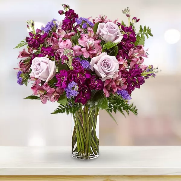 A close-up shot of a clear glass vase filled with purple and pink flowers, including roses, alstroemeria, and other varieties, with visible green stems in the water, placed on a light-colored surface with a blurred background.
