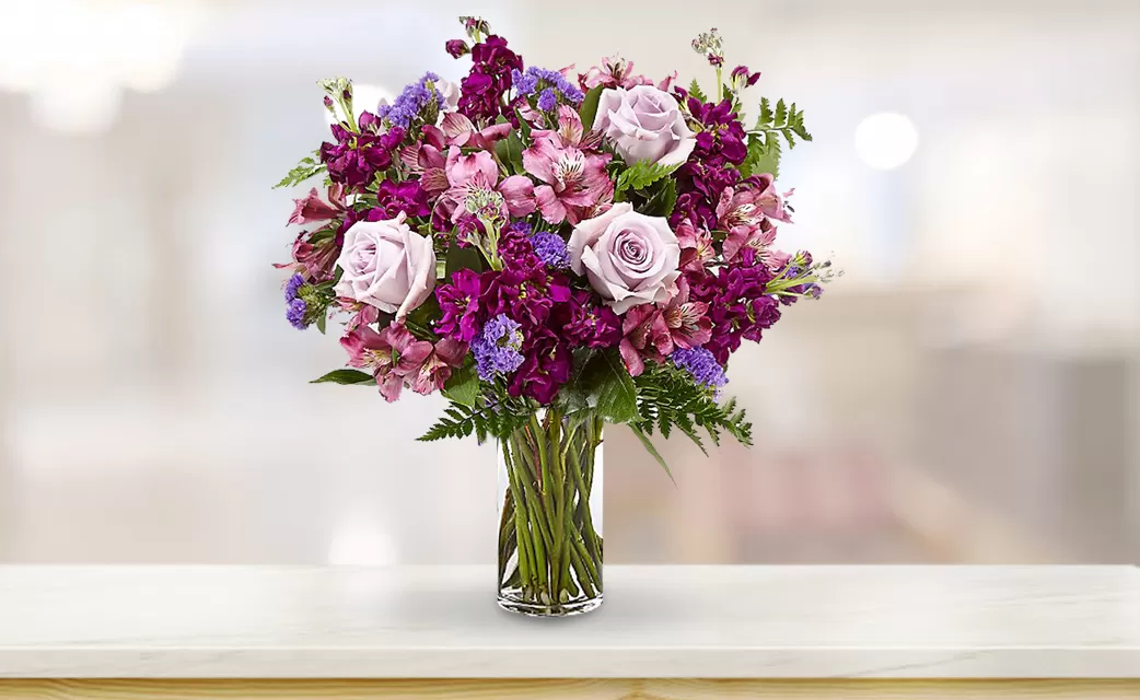 twighlight A close-up shot of a clear glass vase filled with purple and pink flowers, including roses, alstroemeria, and other varieties, with visible green stems in the water, placed on a light-colored surface with a blurred background.