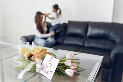 A selective focus shot of a Mother's Day breakfast in bed setup, with a blurred mother and child in the background. A glass-topped table in the foreground holds a plate with a croissant, orange slices, and a glass of orange juice. A small card with "I [heart] MOM" written on it rests against a bouquet of pink and white tulips. The mother, with long brown hair, sits on a black leather couch, facing away from the camera, while a young child with dark hair stands on the couch, interacting with the mother. The room is bright and appears to be a living space.