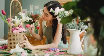 A medium shot of a woman with long dark hair, wearing a gray apron, carefully arranging pink tulips and white lilies in a woven basket and a white pitcher on a wooden table strewn with flower petals and stems.