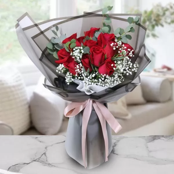 A close-up shot of a bouquet featuring 12 red roses and small white baby's breath flowers, with eucalyptus leaves, wrapped in black and gray paper and tied with a light pink ribbon, placed on a marble surface with a blurred background.