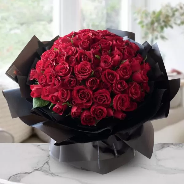 A large bouquet of red roses wrapped in black paper, placed on a marble table with a blurred background of a room with a window.