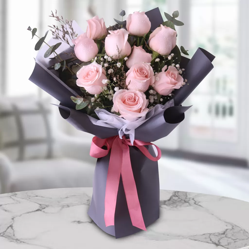 A bouquet of light pink roses with white gypsophila flowers and green eucalyptus leaves, wrapped in dark gray paper and tied with a pink ribbon, placed on a marble table with a blurred background.