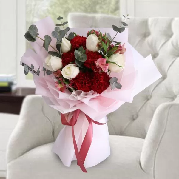A close-up shot of a bouquet featuring red carnations, white roses, and pink alstroemeria flowers, with eucalyptus leaves, wrapped in pink and white paper and tied with a red ribbon, placed on a light-colored armchair with a blurred background.