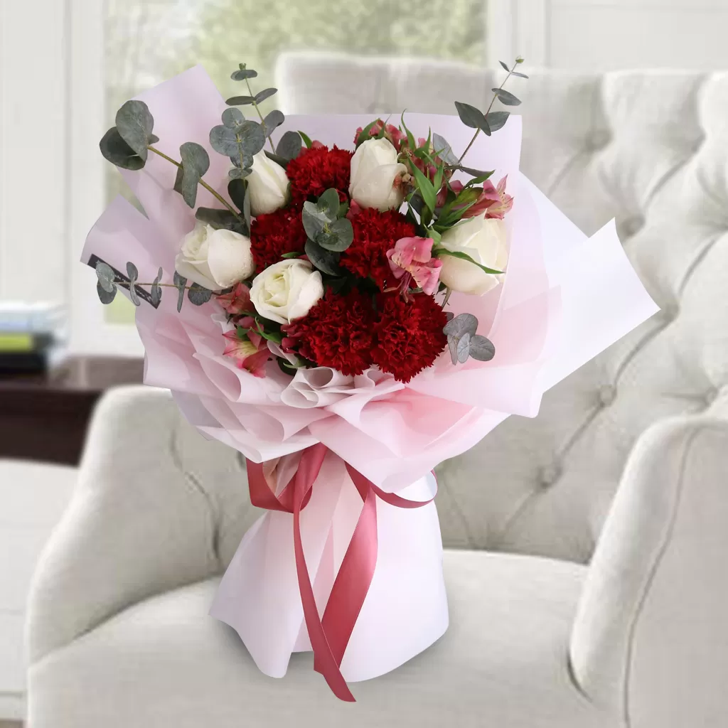 Cara A close-up shot of a bouquet featuring red carnations, white roses, and pink alstroemeria flowers, with eucalyptus leaves, wrapped in pink and white paper and tied with a red ribbon, placed on a light-colored armchair with a blurred background.