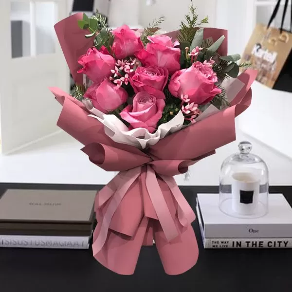 This image shows a bouquet of pink roses with green eucalyptus leaves, wrapped in pink paper and tied with a pink ribbon, placed on a table with two books and a glass cloche with a candle inside. The background is blurred, showing a room with a window and a shopping bag.