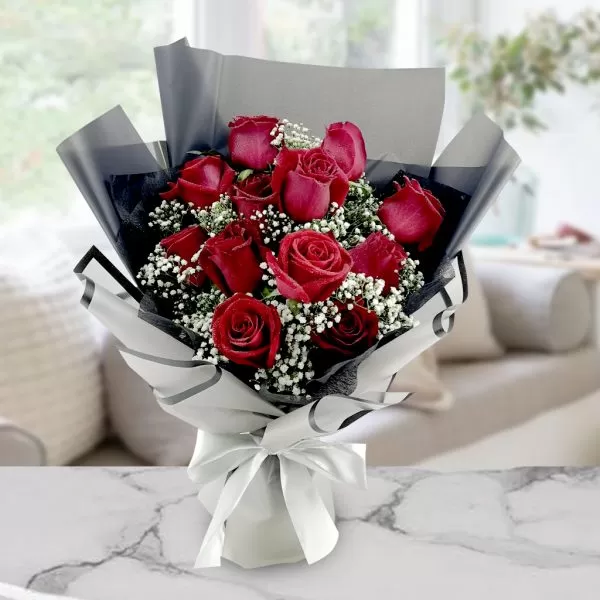 A bouquet of red roses with white baby's breath flowers, wrapped in black and white paper and tied with a white ribbon, placed on a marble table with a blurred background of a room with a window.