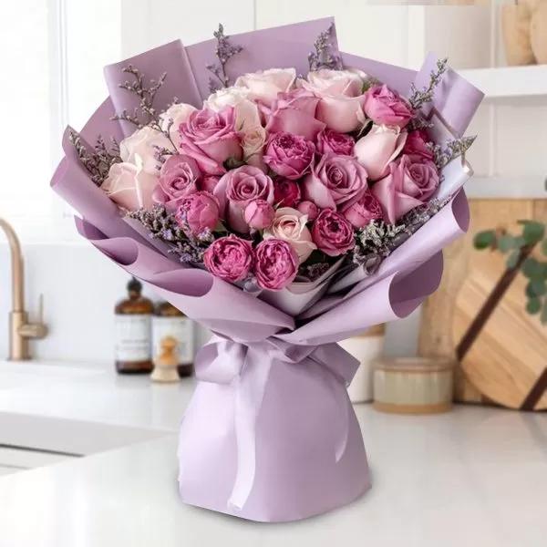 A bouquet of pink and purple roses wrapped in light purple paper and tied with a light purple ribbon, placed on a white countertop with a blurred kitchen background.