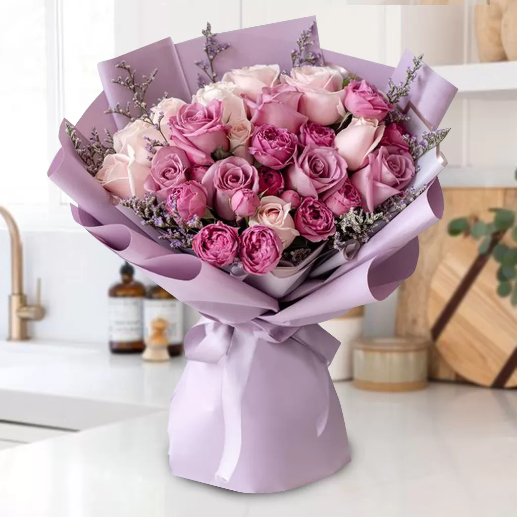 Chic_Pink A bouquet of pink and purple roses wrapped in light purple paper and tied with a light purple ribbon, placed on a white countertop with a blurred kitchen background.